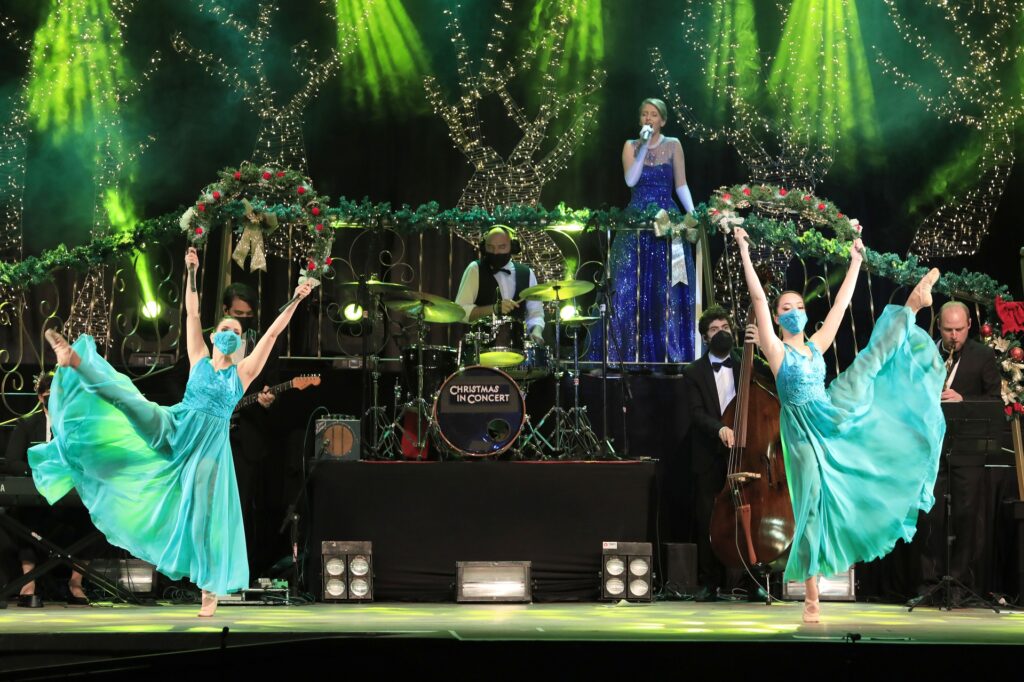 Dançarinas com vestidos turquesa no palco do Christmas in Concert, com banda e cantora ao fundo e iluminação verde no palco