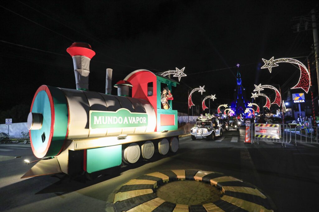 Carro alegórico do Mundo a Vapor, simulando uma locomotiva de trem, verde e vermelha, na Parada de Natal. Ao fundo, a Catedral de Pedra iluminada