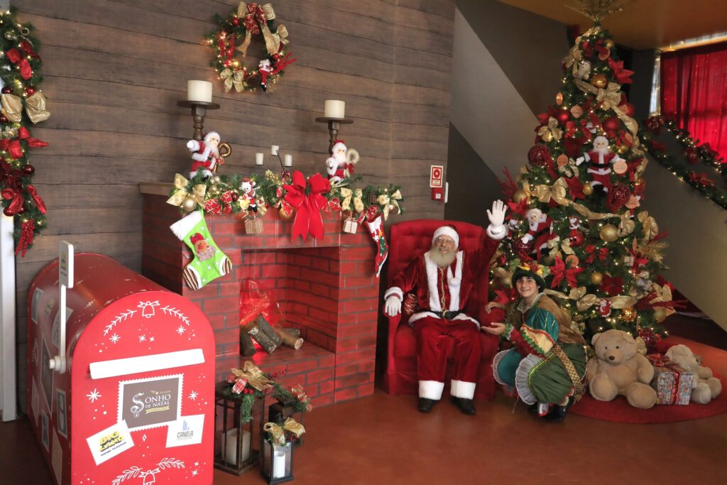 Papai Noel em um ambiente com decoração natalina e um pinheiro alto decorado montado ao fundo