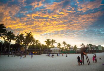 Pôr do sol em Praia de Porto de Dunas, em Aquiraz. Pessoas jogam futebol na areia e o sol reflete luz amarela nas nuvens.
